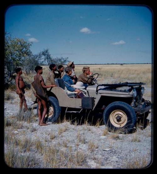 Expedition: John Marshall holding a camera, with Lorna Marshall, Ngani, and other expedition members riding in Jeep with him; two men with hunting equipment are standing nearby