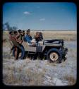 Expedition: John Marshall holding a camera, with Lorna Marshall, Ngani, and other expedition members riding in Jeep with him; two men with hunting equipment are standing nearby
