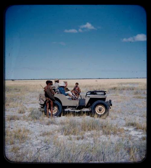 Expedition: John Marshall holding a camera, with Lorna Marshall, Ngani, and other expedition members riding in Jeep with him; two men with hunting equipment are standing nearby