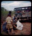 Expedition: John Marshall sticking his head through a flat tire, with other men standing nearby; another man is fixing an expedition truck in the background, on their trip from "Kubi"