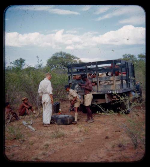 Expedition: John Marshall, Ngani, and another man standing and looking down at a flat tire on the ground, with other people in the background, on their trip from "Kubi"