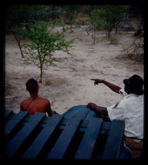 Expedition: Thoria sitting on a truck fender and pointing at something, with another man sitting next to him, seen from behind