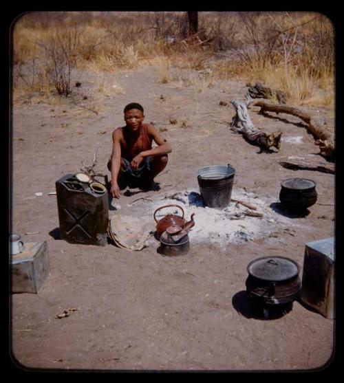 Expedition: Man wearing a ragged jersey and squatting at extinguished fire for heating water to wash at the expedition camp