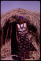 N!ai wearing Western clothing, standing in front of a hut