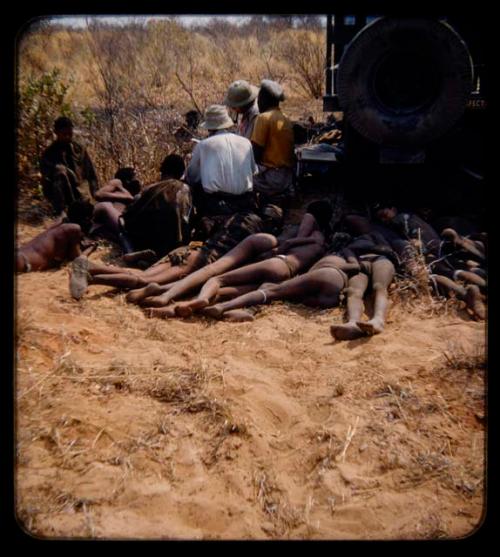 Expedition: J. O. Brew interviewing a man near a truck, assisted by two interpreters, with a group of people lying on the ground in the foreground