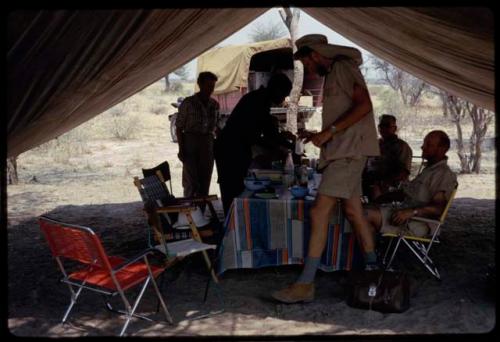 Expedition members gathered inside a tent (including O.P.M. Prozesky, C.J. Mathias, Wulf Haacke, and Lorna Marshall) and Philip Hameva serving food