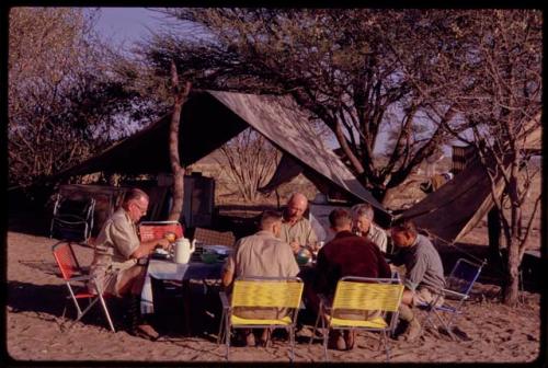 C.J. Mathias, O.P.M. Prozesky, Lorna Marshall, Kurt Ahrens, Nicholas England, and Wulf Haack sitting and eating at the expedition camp breakfast table