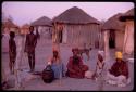 Women sitting in front of huts, with a couple of boys standing next to them