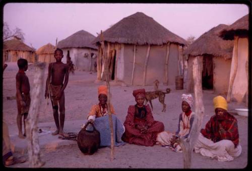 Women sitting in front of huts, with a couple of boys standing next to them