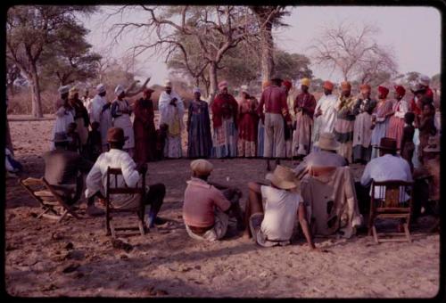 Kandyendye in a pink shirt singing, with a group of women wearing "modern" Herero dress and men sitting in chairs