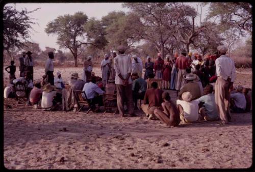 Kandyendye in a pink shirt singing, with a group of women wearing modern Herero dress and men sitting in chairs