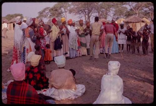 Man and Kandyendye in a pink shirt singing for a group of women wearing modern Herero dress and children