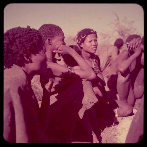 Groups, "Nuclear Family": Group of people sitting by "/Gao Music's" skerm, including Khuan//a ("Gao Medicine's" sister) wearing copper rings in her hair, //Khuga with her hand to her face and /Naoka ("Gao Medicine's" first wife)