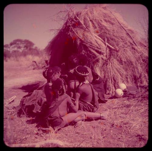 Groups, "Nuclear Family": Group of women and girls sitting in front of a skerm, with an assegai leaning against the skerm and a quiver hanging on it