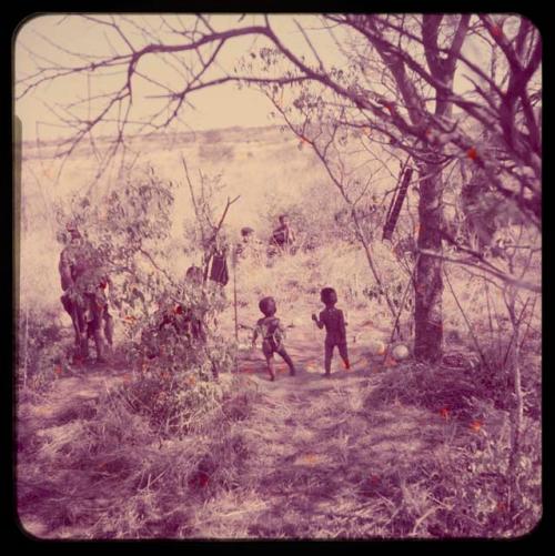 Skerm: Two children standing in "Gao Helmet's" dwelling place, with people sitting in the background