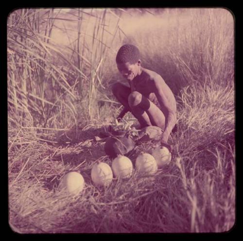 Water: ≠Gao (leader of Band 10) filling ostrich egg shells with water at a waterhole, with five ostrich egg shells with stoppers and an Ovambo pot on the ground next to him