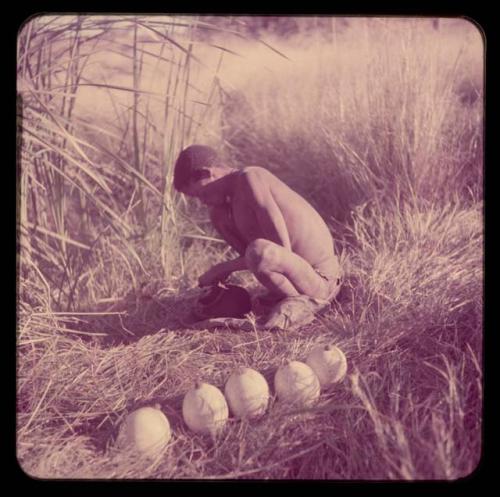 Water: ≠Gao (leader of Band 10) filling an Ovambo pot with water at a waterhole, with five ostrich egg shells with stoppers on the ground next to him