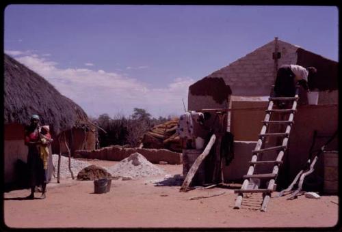 People working on a new house, with a woman holding a baby standing near them, in the compound of Boy Moapane, brother of the Kgalagari chief