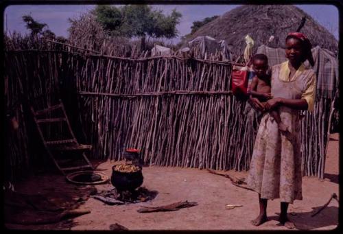 Woman standing, holding a baby at a cooking place inside the fence of her kraal