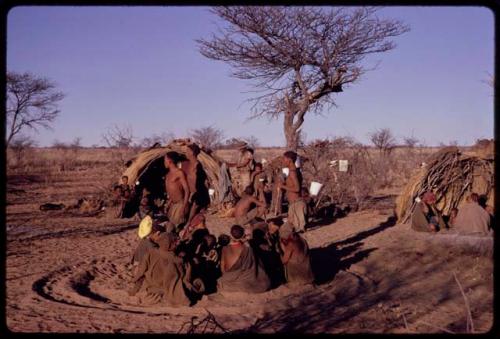 Men performing a curing dance around a circle of seated women, with people sitting and standing next to a skerm in the background