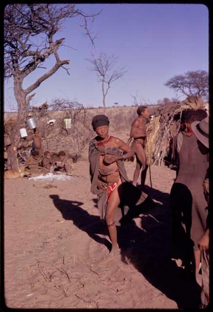 /Oβa (wife of !Kxam) standing and holding her breast at a social dance, with a group of children sitting in the background