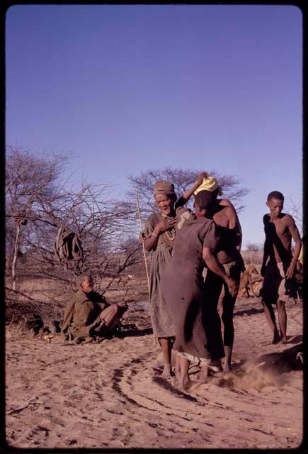 Group of people performing a social dance (including N//aba giving her kerchief to /Xoxa and Sãõ?//e beside N//aba), with "Old Xama" from Werft 2 sitting near them