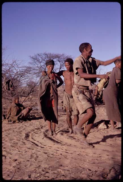 Man wearing shoes and performing a social dance, with /Gaishay and /Oβa behind him