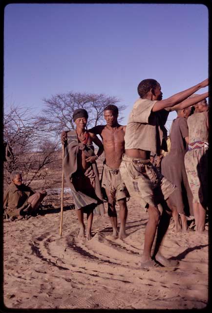 Man wearing a shirt, shorts and shoes and performing a social dance, with /Oβa and /Gaishay behind him and "Old Xama" (from Werft 2) sitting in the background