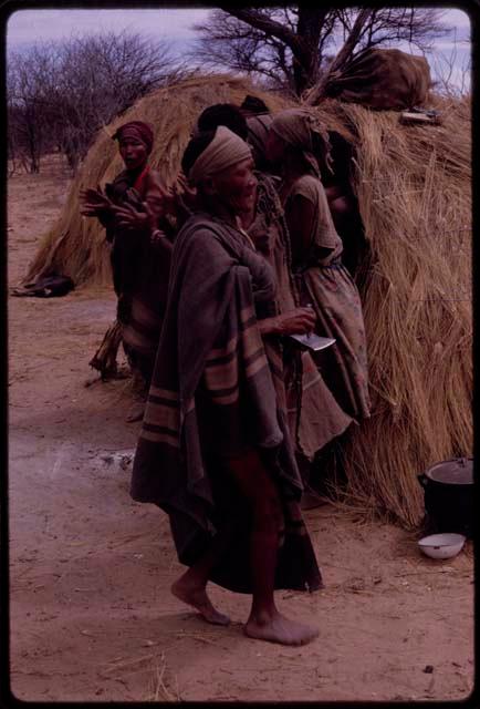 Sãõ?//e playing an axe blade, with women standing and clapping behind her, near a skerm used for the menstruating girl in the Eland Dance