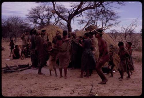 Group of people performing the Eland Dance in front of a skerm, including !Kxam wearing horns on his head