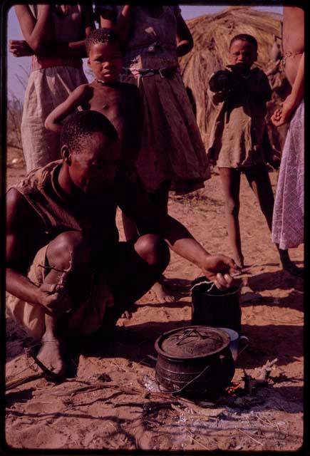 Man squatting and cooking, with other people standing behind him