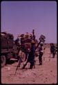 Men standing near a road sign that points to Maun in one direction and Ghanzi in the other, with inspectors examining expedition trucks in the background