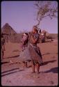 Women, including one woman holding a baby, in a village, with huts in the background