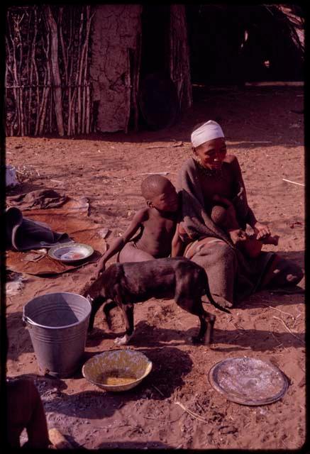 Buhu sitting in front her skerm and her child leaning against her, with a dog in the foreground