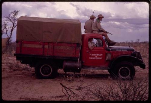 O.P.M. Prozesky and Wulf Haacke in profile, holding a gun and sitting on top of expedition Dodge that reads "Peabody Museum of Harvard University," on the way to Ghanzi District