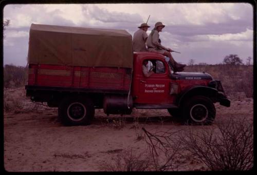 O.P.M. Prozesky and Wulf Haacke in profile, holding a gun and sitting on top of expedition Dodge that reads "Peabody Museum of Harvard University," on the way to Ghanzi District