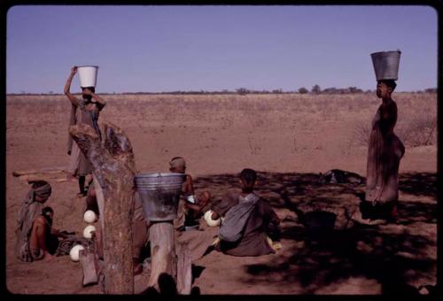 Women holding their buckets on their heads while others sit and fill ostrich eggshells with water