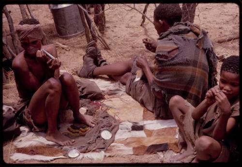 Dadum sitting on animal skins and smoking while her husband (Tama) sews with a boy sitting behind him