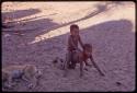 Child riding on another's back in the sand, with a dog asleep beside them