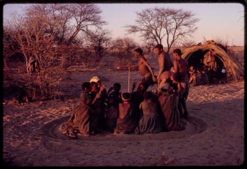 /Xoxa, /Gaishay, and /'O/hei/ performing a curing dance around a group of women sitting in a circle and clapping, with other people sitting in front of a skerm in the background
