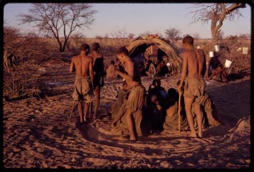 /Xoxa, /Gaishay, /'O/hei/, and Tama performing a curing dance around a group of women sitting in a circle, with other people sitting in front of a skerm in the background