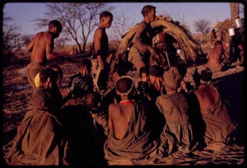 Group of women sitting in a circle and clapping as three men (/Xoxa, /Gaishay, and /'O/hei/) perform a curing dance