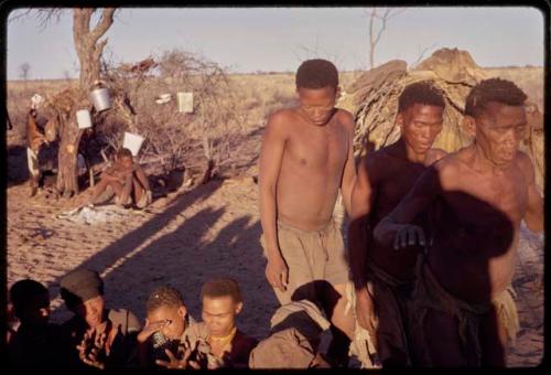 /Xoxa, /Gaishay, and Tama performing a curing dance around a group of women sitting in a circle; the shadow of a photographer is visible