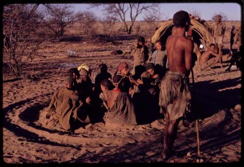 Group of women sitting in a circle for a curing dance and /Gaishay standing nearby, with other people in the background