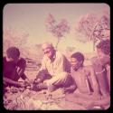 Expedition: Gau fixing Laurence Marshall's shoe by making a hole with an awl and sticking thread through the hole, with "Gao Medicine" and ≠Toma sitting with them