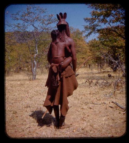 Woman carrying a child on her back and holding a stick in her hand, seen from behind