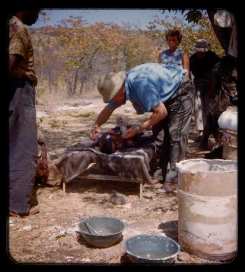 Boy lying on a cot, Eric Williams making a cast of his face; people standing nearby and watching