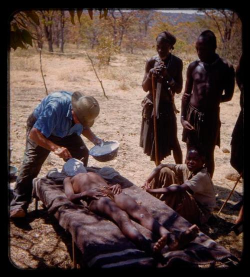 Boy lying on a cot, Eric Williams making a cast of his face; people standing nearby and watching