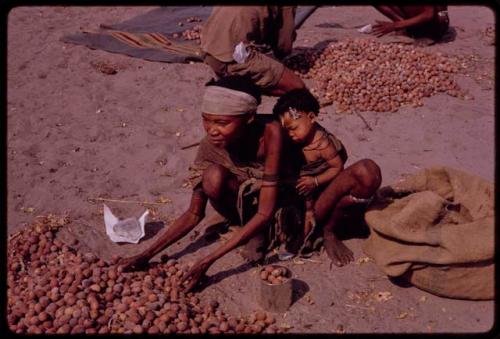 //Khuga, "Crooked /Qui"'s wife, and their new baby crouching beside her pile of mangetti nuts