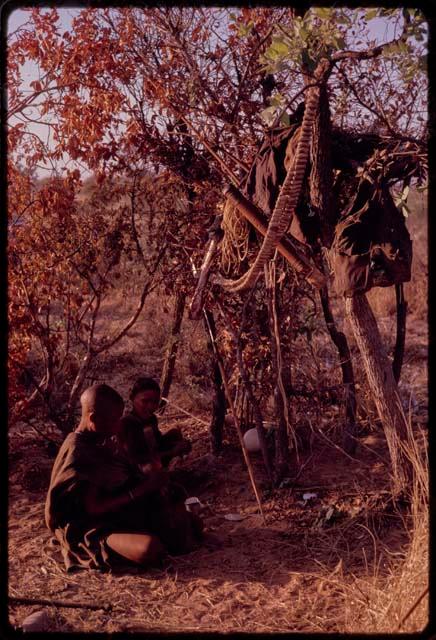 Two women sitting by a tree where their belongings hang